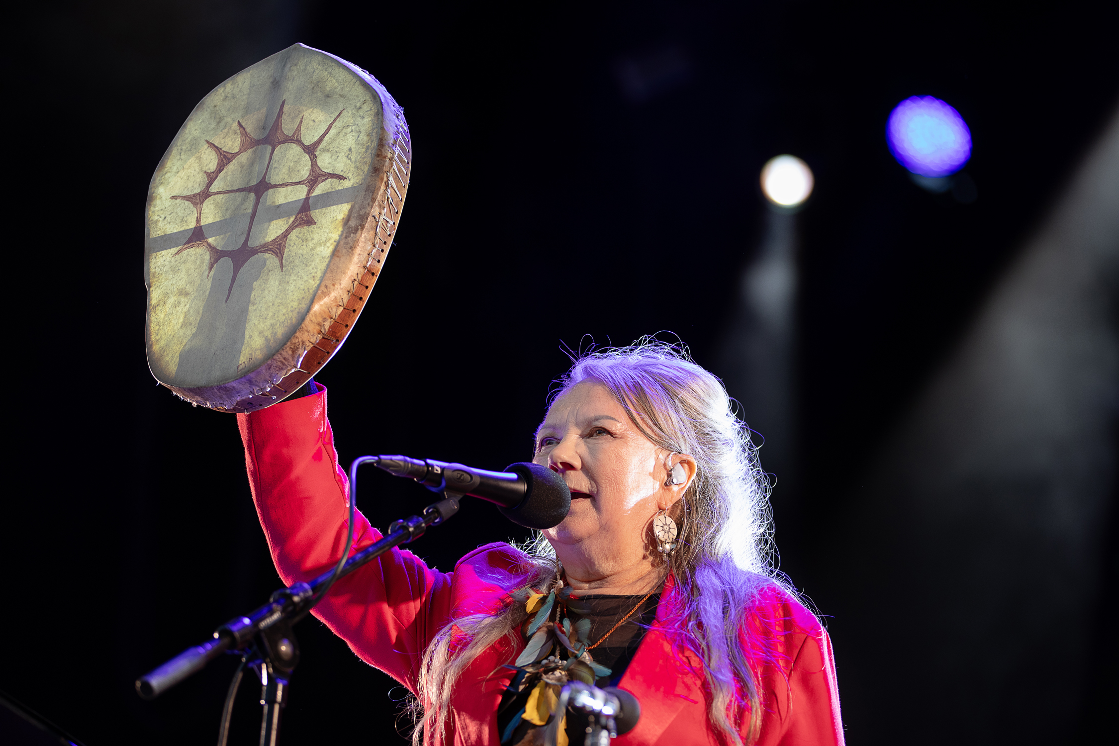 Mari Boine holder opp en samisk tromme mens hun synger inn i mikrofonen på scenen.